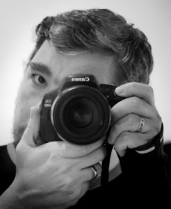 Self portrait, black and white. Dark hair, headshot, face obscured by a canon camera being held up to one eye.